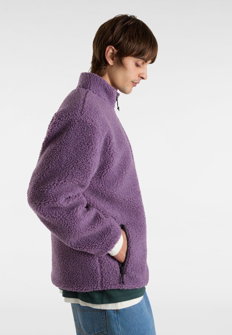 Lila Fleecejacke mit hohem Kragen, seitlichen Taschen und strukturierter Oberfläche. Über einem grün-weißen Schichthemd getragen.