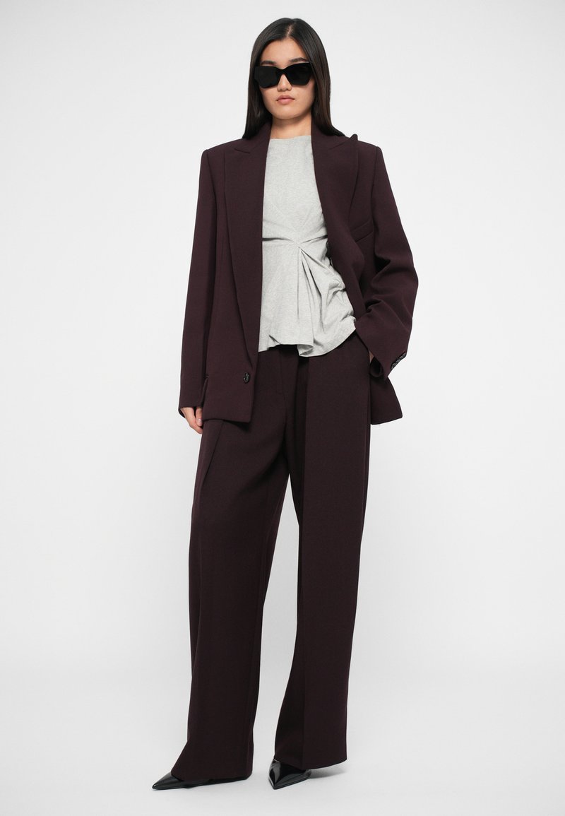 Blazer sartorial bordeaux sopra un top grigio chiaro plissettato, abbinato a pantaloni a gamba larga e tacchi a punta neri. Gli occhiali da sole neri completano il look.