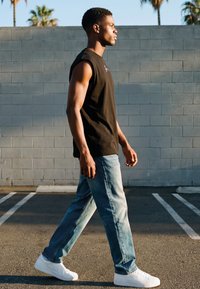 Ung mann som går utendørs iført svart ermeløs skjorte, blå jeans og hvite joggesko ved en grå murvegg.