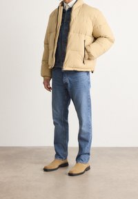 Beige puhvärjakke med lynlås, blå denimskjorte under, blå jeans og lys brune slip-on sko på et teksturert gulv. Minimalistisk design.