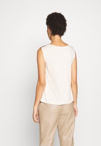Top senza maniche di un beige chiaro con scollatura arrotondata, dotato di dettagli decorativi sulle spalle e realizzato in un tessuto morbido e liscio.