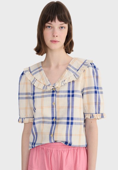 Jeune femme aux cheveux bruns courts portant un chemisier crème à carreaux bleus et oranges, col à volants, et jupe taille haute rose.