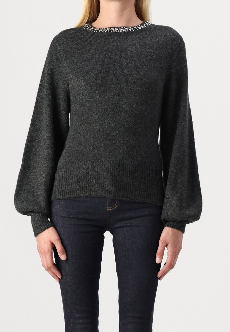 Femme portant un pull gris foncé à manches ballon et col orné, associé à un jean bleu foncé ajusté.