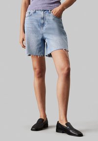 Pantalones cortos de mezclilla azul claro con dobladillo deshilachado, diseño de cinco bolsillos y presillas para cinturón, combinados con zapatos negros sin cordones.