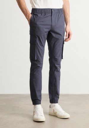 Homme portant un pantalon cargo bleu marine avec des ourlets élastiques et des poches, assorti à des chaussettes blanches et des baskets blanches sur un sol uni.