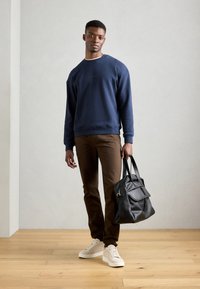 Navy sweatshirt, brown pants, and white sneakers. Holding a black tote bag with straps. Floor is wood, walls are light gray.