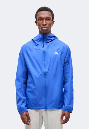 Un homme est de face, vêtu d’une veste à capuche bleu vif zippée et d’un pantalon gris clair, sur un fond uni.