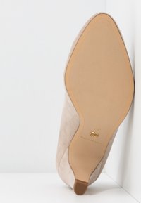Chaussure à talon haut pour femme en daim beige reposant sur le côté, mettant en valeur la semelle propre et l’extrémité carrée du talon sur fond blanc.