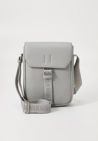 Bolso cruzado gris de cuero sintético con cierre de solapa, con un broche plateado y una correa de tela ajustable con el grabado "ck".