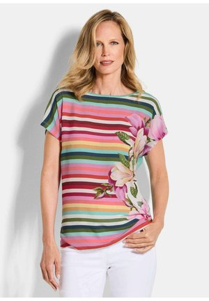 Femme aux cheveux blonds portant un haut à manches courtes avec des rayures horizontales colorées et un grand imprimé de fleur rose, assorti à un pantalon blanc.