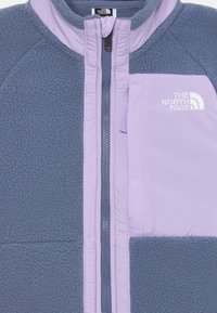Plava flis jakna s lavandom zatvaračem i džepom na prsima, s izvezenim logotipom The North Face u bijeloj boji.