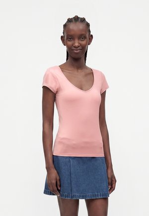 Femme aux cheveux tressés portant un haut rose à manches courtes avec col en V et une jupe en jean bleue, debout devant un fond clair uni.