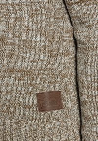 Maglione lavorato a maglia con un motivo marrone e crema, dalla superficie testurizzata. Un'etichetta in pelle marrone è cucita sul lato inferiore.