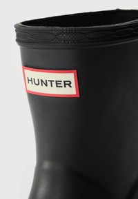 Botte noire en caoutchouc montante avec un bord supérieur texturé et une étiquette rouge et blanche de marque avec le mot "HUNTER". Surface lisse.