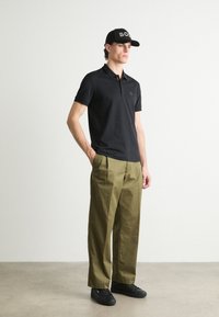 Black polo shirt with short sleeves and a collar, paired with olive green loose-fit trousers and black sneakers, complemented by a black cap.