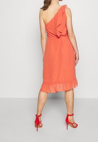 Koralowa sukienka na jedno ramię z falbaną i falbaniastym dołem, wykonana z lekkiego materiału. Dobranie do niej czerwone sandały na wysokim obcasie.