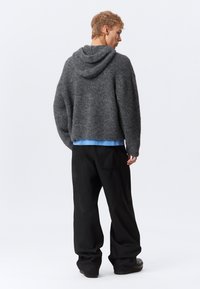 Joven vestido con un suéter gris holgado con capucha, camisa azul claro, pantalones anchos negros y zapatos negros, de pie de espaldas contra un fondo blanco.