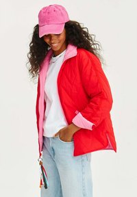 Červená a ružová prešívaná bunda s vysokým golierom, biela tričko, svetlo modré džínsy a ružová čiapka, doplnené farebným kľúčenkom.