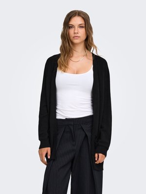 Mujer con cabello ondulado castaño claro lleva un cárdigan negro, camiseta blanca sin mangas y pantalones anchos de rayas negras sobre un fondo claro y liso.