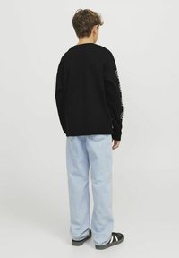 Camisa negra de manga larga con detalles impresos en blanco en las mangas, combinada con jeans holgados de color azul claro y zapatillas negras, vista desde atrás.