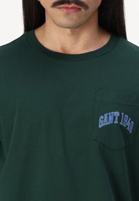 Grøn bomulds T-shirt med korte ærmer. Har en rektangulær lomme på venstre bryst med blå "GANT 1949" broderi detalje.