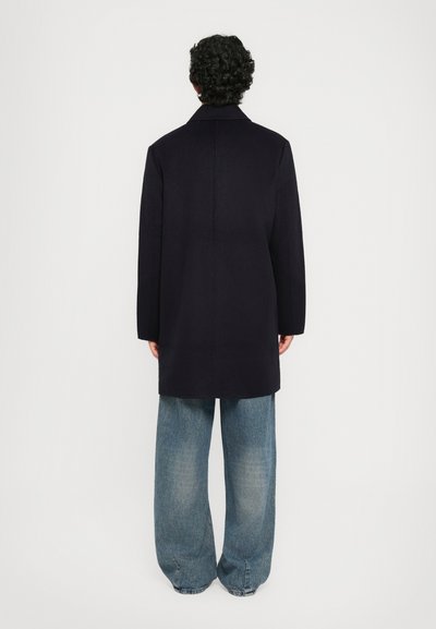 Man with curly dark hair wearing a black coat and loose blue jeans, standing facing a plain white background.