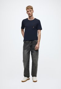 T-shirt a maniche corte di colore blu navy con taschino e logo giallo, abbinata a jeans neri sbiaditi e sneakers bianche con suole marroni.