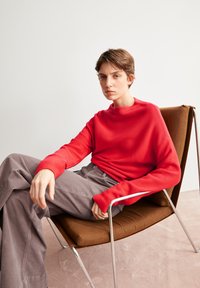 Rode, lange mouwen, hooghalstrui in een gestructureerde knit. Grijze corduroybroek. Moderne stoel met een bruine kussen en een metalen frame. Neutrale achtergrond.