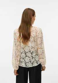 Koronkowa bluzka z długim rękawem, zdobiona kwiatowym wzorem, w kolorze kremowym, o lekko prześwitującej fakturze. Zaokrąglony dół i marszczone mankiety.