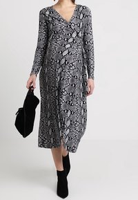 Kobieta ubrana w midi sukienkę z długim rękawem w czarno-biały wzór w węża, trzymająca czarną torebkę i nosząca czarne botki za kostkę.