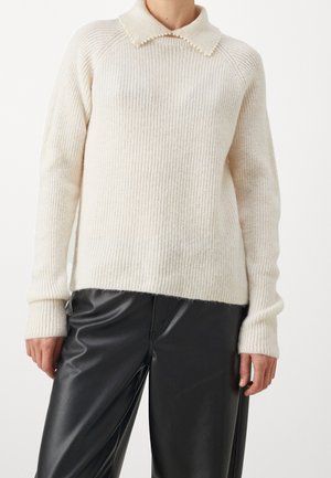 Personne portant un pull côtelé crème avec un col pointu et une bordure perlée, associé à un pantalon en cuir noir, debout devant un fond blanc.