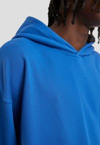 Sudadera con capucha azul de textura suave, corte relajado y escote en V profundo por encima de la capucha. Detalle de costura visible en los hombros.