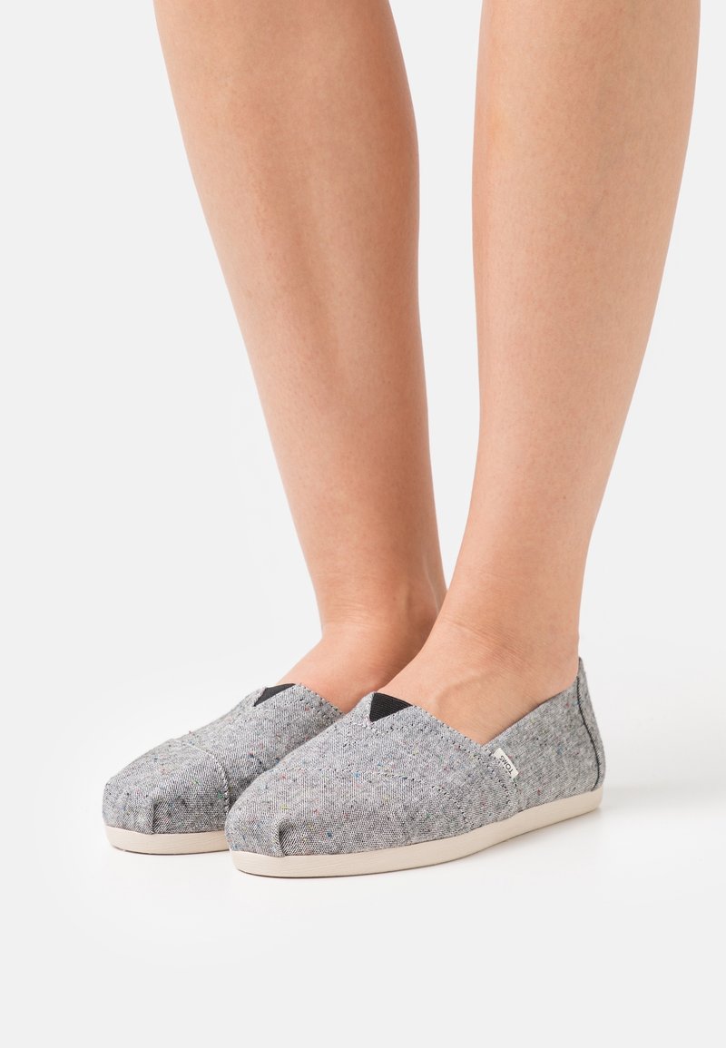 Des chaussures décontractées slip-on gris clair avec des éclats multicolores subtils, portées pieds nus sur fond blanc.