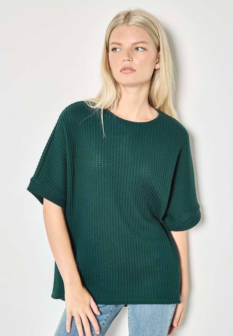Dunkelgrünes Strickoberteil mit kurzen Ärmeln, lockerem Schnitt, rundem Ausschnitt und strukturierter, gerippter Stoffstruktur. Kombiniert mit hellblauen Jeans.