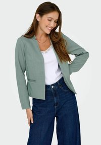 Femme souriante aux longs cheveux bruns portant un blazer vert clair, un haut blanc orné de dentelle et un jean taille haute bleu foncé, sur un fond blanc.