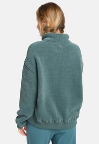 Sudadera de felpa en color teal de gran tamaño con textura acanalada, cuello embudo y dobladillo y puños elásticos, que presenta un pequeño logo en la parte posterior.