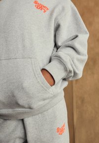 Sudadera gris con puños acanalados y un bolsillo frontal. Presenta un logo naranja de "Superdry" en el pecho y una textura suave.