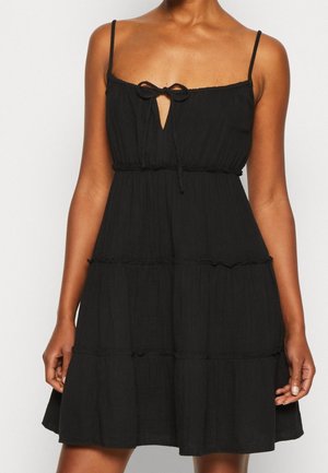 Robe noire sans manches avec bretelles spaghetti réglables, un nœud au niveau du col et un ourlet à volants en plusieurs couches, fabriquée en tissu léger.