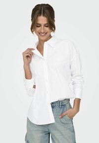 Camicia bianca con collo a cappotto, maniche lunghe e vestibilità rilassata, abbinata a pantaloni corti di jeans blu chiaro.