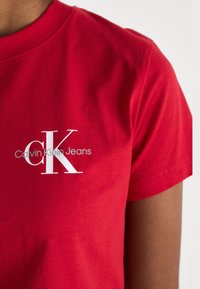 Rdeča bombažna majica s kratkimi rokavi z belim logotipom "Calvin Klein Jeans" in izrazitim dizajnom "CK" na prsih. Klasičen kroj.