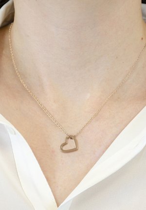 Gold-toned necklace with a heart-shaped pendant, featuring a delicate chain and smooth metallic texture, worn over a white blouse.