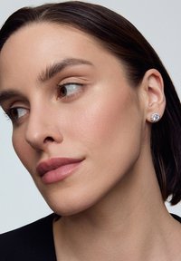 Okrągłe srebrne kolczyki z kwiatowym wzorem, ozdobione wieloma małymi przezroczystymi kamieniami w skupisku, noszone przez modelkę o neutralnym wyrazie twarzy.