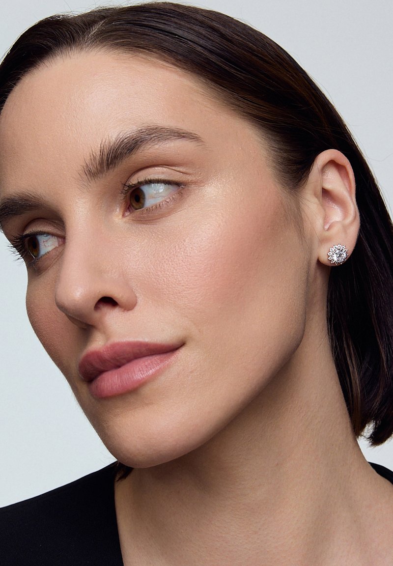 Okrągłe srebrne kolczyki z kwiatowym wzorem, ozdobione wieloma małymi przezroczystymi kamieniami w skupisku, noszone przez modelkę o neutralnym wyrazie twarzy.