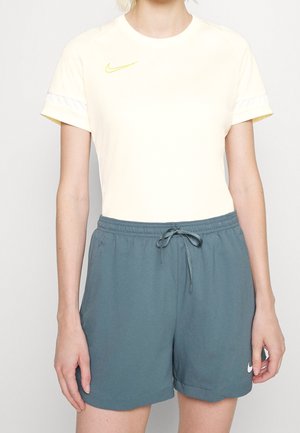 Osoba ubrana w bladożółtą koszulkę Nike i niebieskie spodnie Nike z troczkami, stojąca na jednolitym jasnym tle.