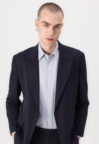 Marineblauwe, op maat gemaakte blazer met notch-lapels en dubbele knopen, gecombineerd met een lichtblauwe overhemd met kraag; gladde stof, gestructureerd ontwerp, frisse uitstraling.
