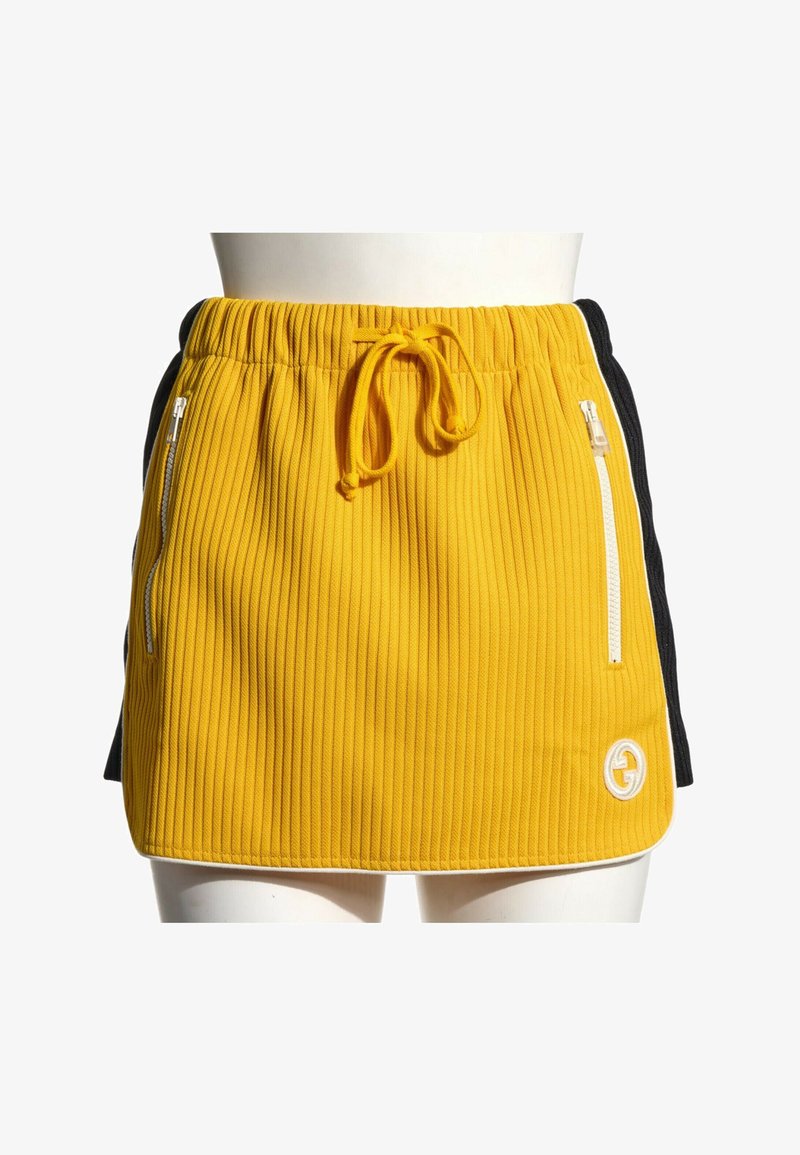 Mini-jupe côtelée jaune avec poches zippées blanches, taille à cordon et panneaux latéraux noirs sur un mannequin.