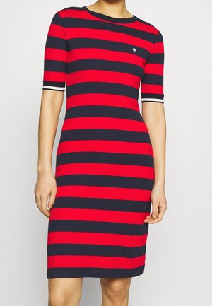 Kobieta ubrana w dopasowaną sukienkę do kolan z poziomymi czerwonymi i granatowymi paskami oraz krótkimi rękawami z białym wykończeniem.