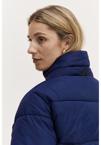 Marinblå puffer-jacka med hög krage, kviltat mönster och svart logotypdetalj på baksidan. Slät, glansig yta.