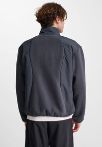 Mörkgrå fleecejacka med ståkrage, elastisk nederkant och kontrasterande paneler. Slät yta med matt finish, designad för en avslappnad passform.