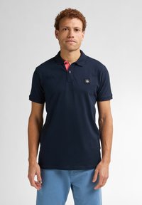 Mörkblå polo-t-shirt med en rosa detalj vid kragen, korta ärmar och en logotyppatch på vänster bröst. Slätt bomullsmaterial.